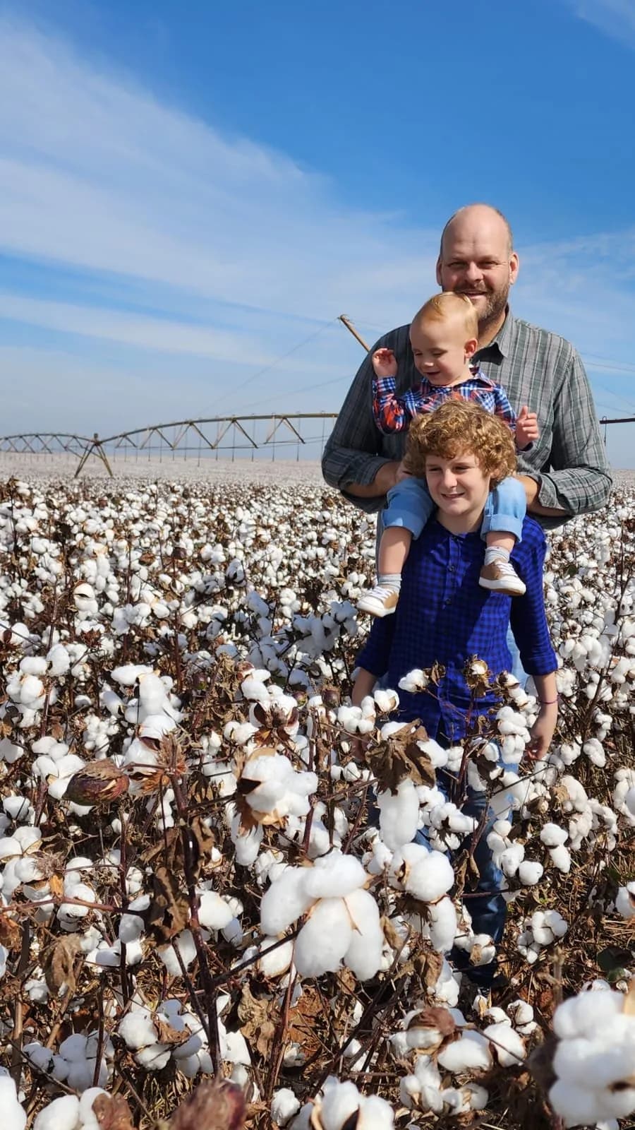 Pai e filhos em um algodoal sob céu azul, com pivô de irrigação ao fundo
